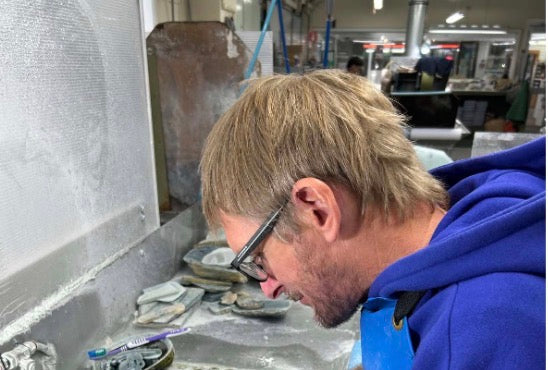 A man with blond hair and glasses, wearing a blue hoodie and blue apron, looking down at pottery pieces on a workbench in a studio.